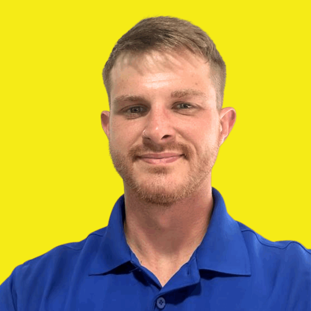 A man with short light brown hair and a trimmed beard, wearing a blue collared shirt, is smiling in front of a bright yellow background.