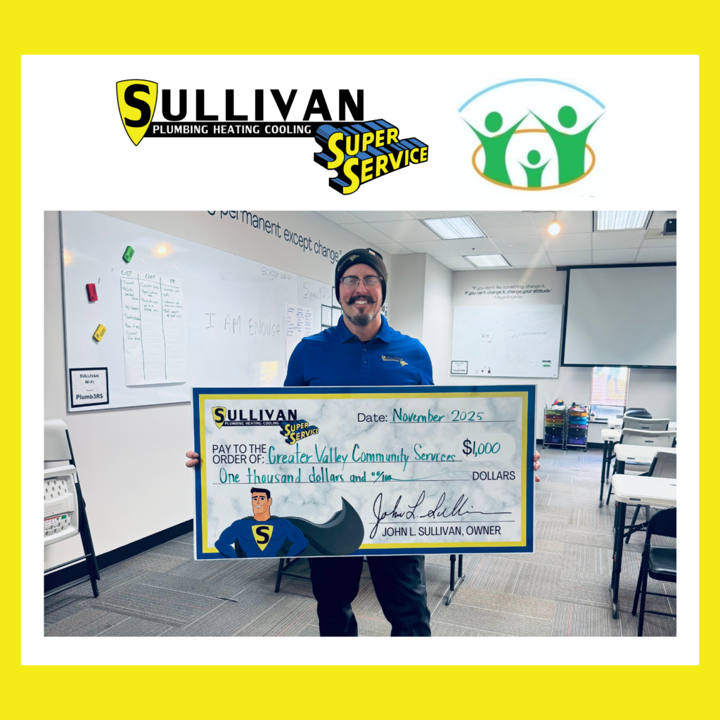 A man stands in a classroom holding a large check for $1,000 from Sullivan Plumbing Heating Cooling, made out to Greater Valley Community Service.