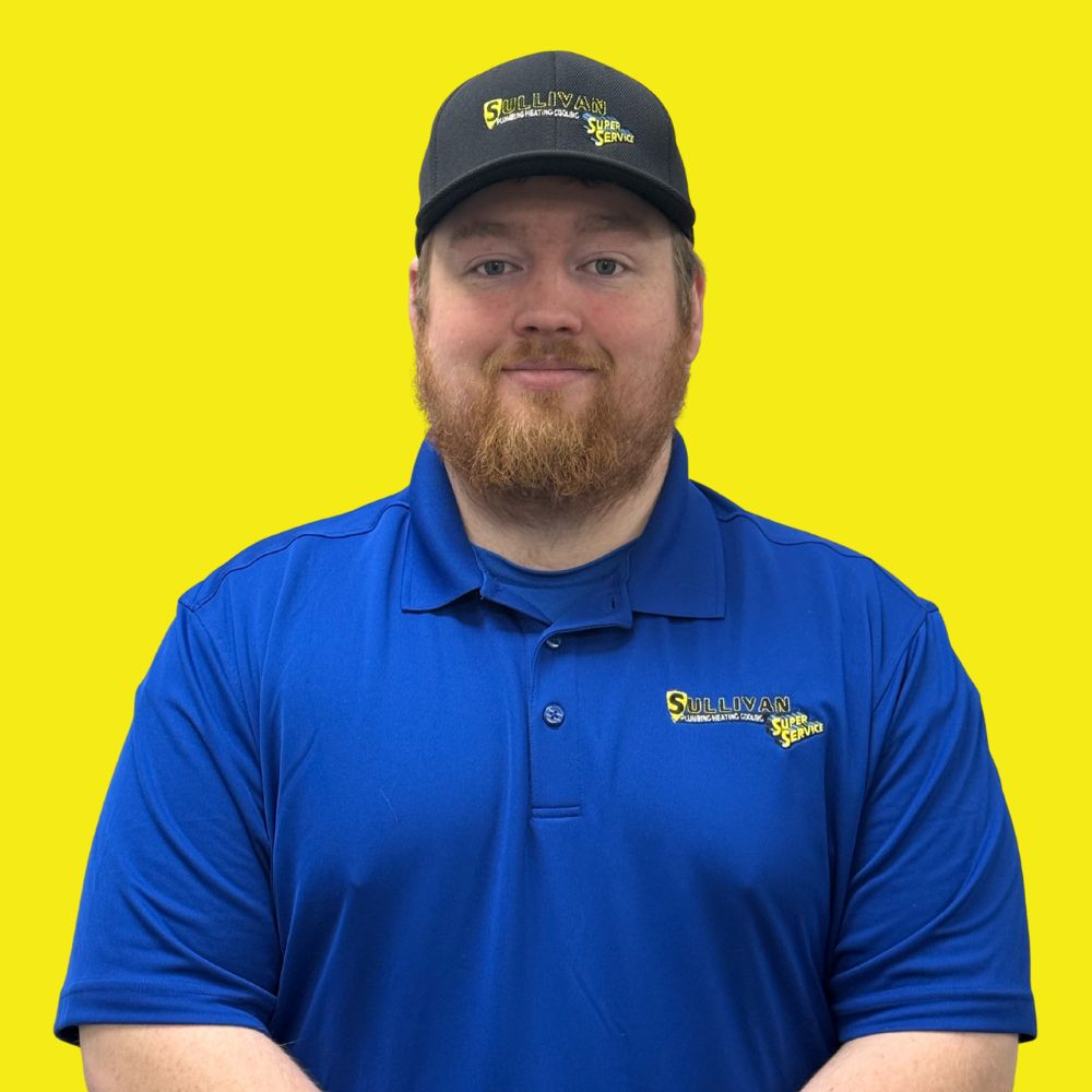 A man with a ginger beard wears a black cap and blue polo shirt with "Sullivan Tire and Auto Service" logos, standing against a bright yellow background.