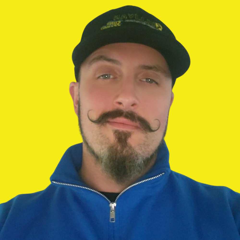 A man with a styled mustache and goatee, wearing a black cap and blue zip-up jacket, poses in front of a plain yellow background.
