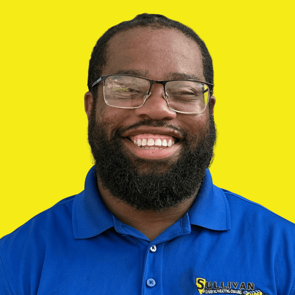 A man with glasses and a beard smiles at the camera, wearing a blue polo shirt with a “Sullivan” logo, against a solid yellow background.