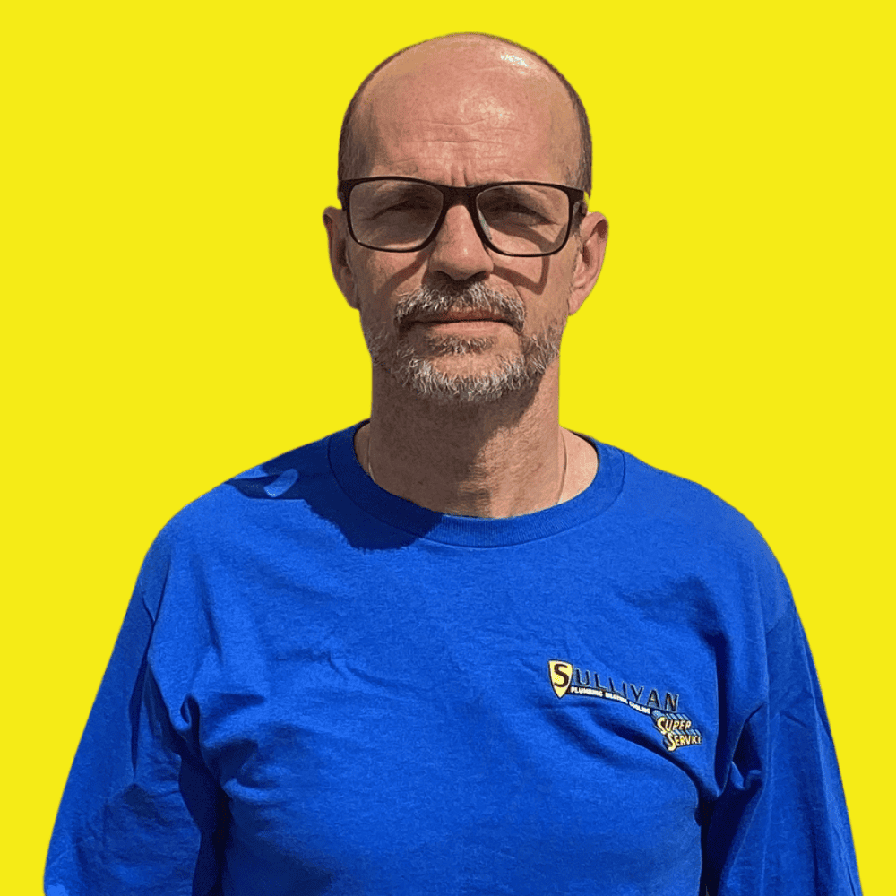 A man wearing glasses and a bright blue “Sullivan Super Service” shirt stands in front of a solid yellow background.