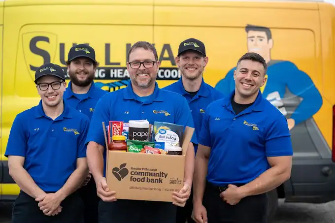 Five men in blue uniforms stand in front of a yellow van, one holding a box of donated food labeled “Greater Pittsburgh Community Food Bank.”.